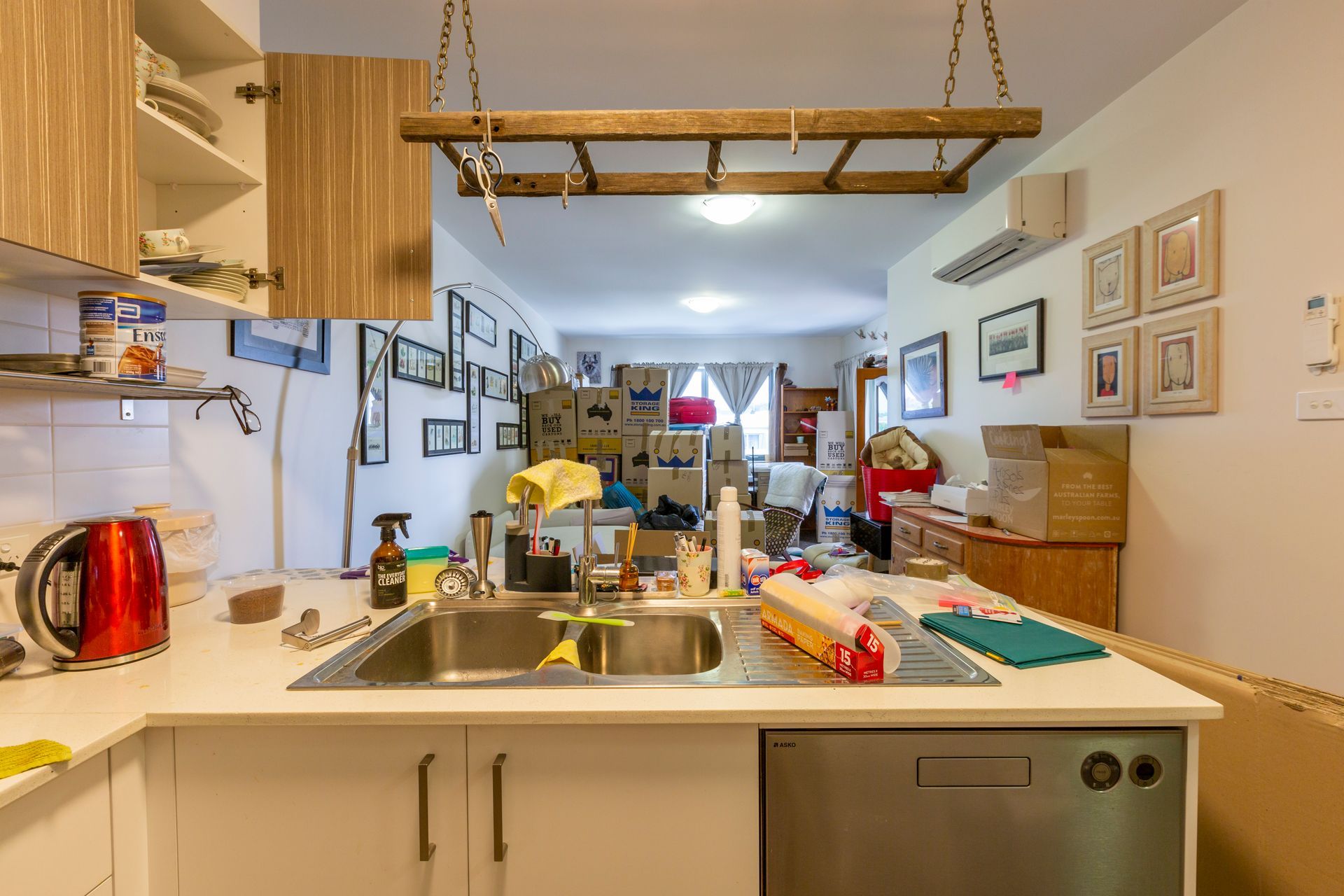 Kitchen with open cabinets, a sink, and a view into a messy living room with boxes and furniture.