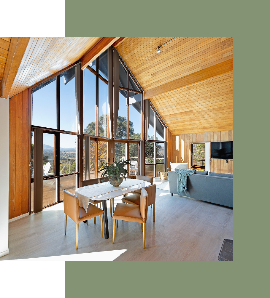 Dining room with large windows, wooden ceiling, table, chairs, and a gray sofa.