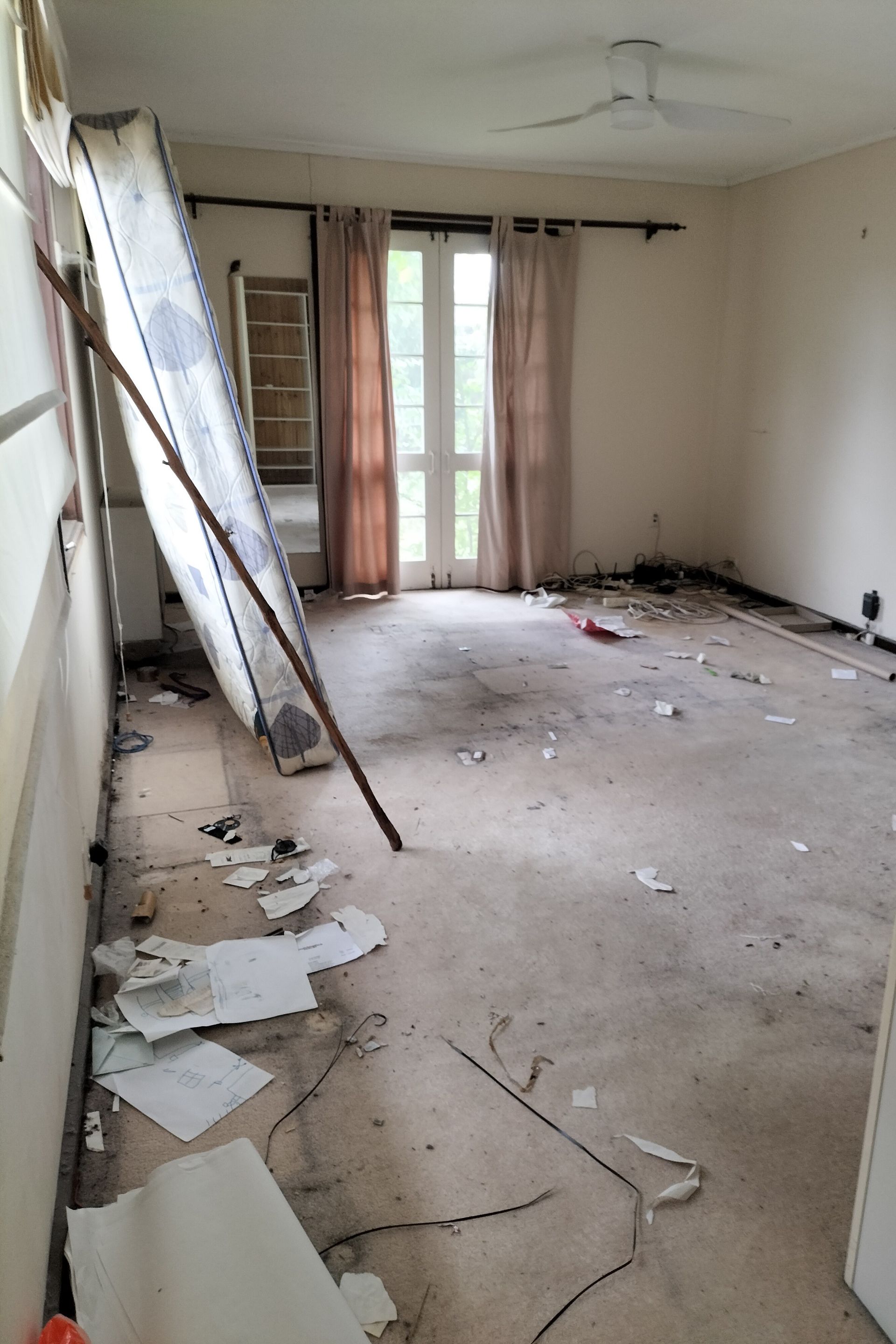 Messy bedroom with wardrobe, dresser covered in clothes, fan, and cluttered floor.