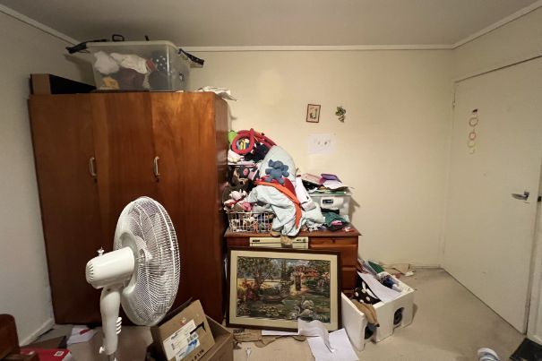 Messy bedroom with wardrobe, dresser covered in clothes, fan, and cluttered floor.