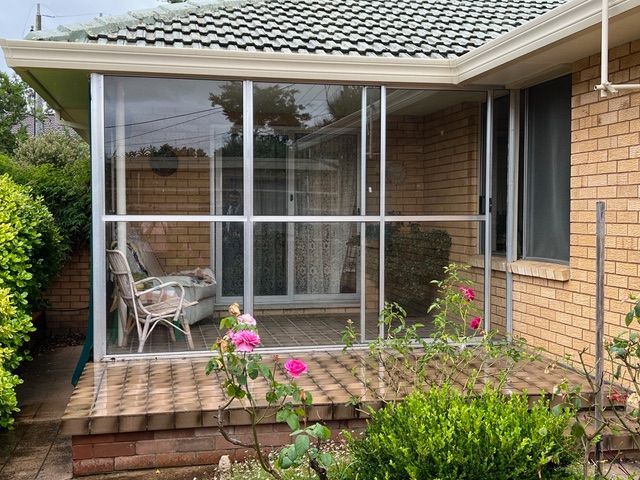 Enclosed patio with aluminum frame and screen, attached to a brick house. Chairs visible inside.
