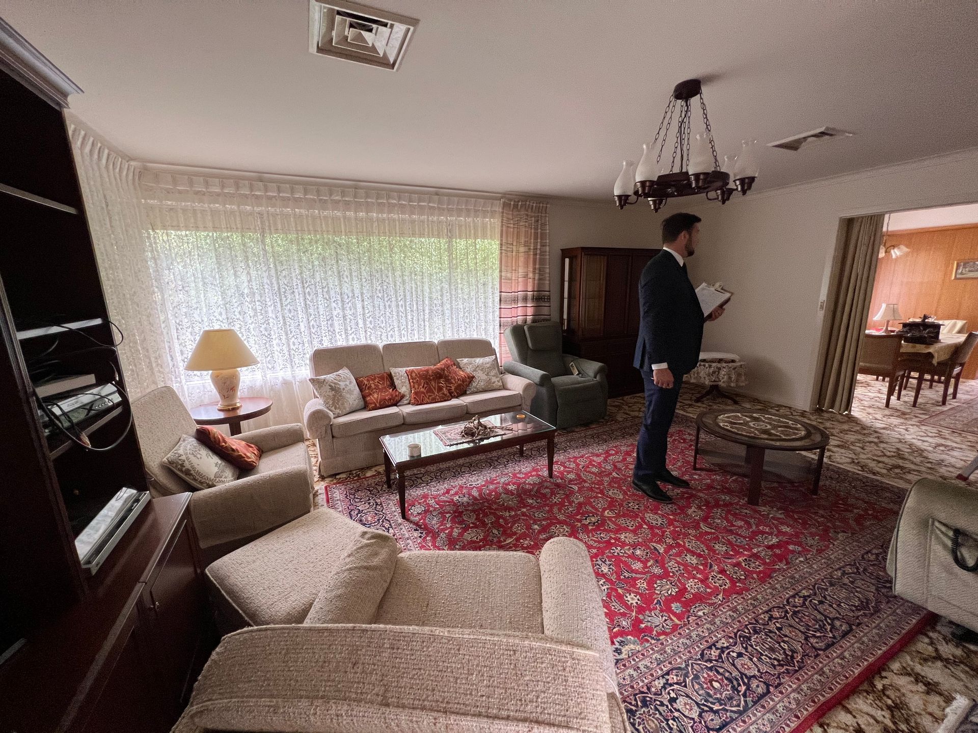 A person in a suit stands in a living room, looking at a document. The room has a rug, sofas, and a dining area visible.