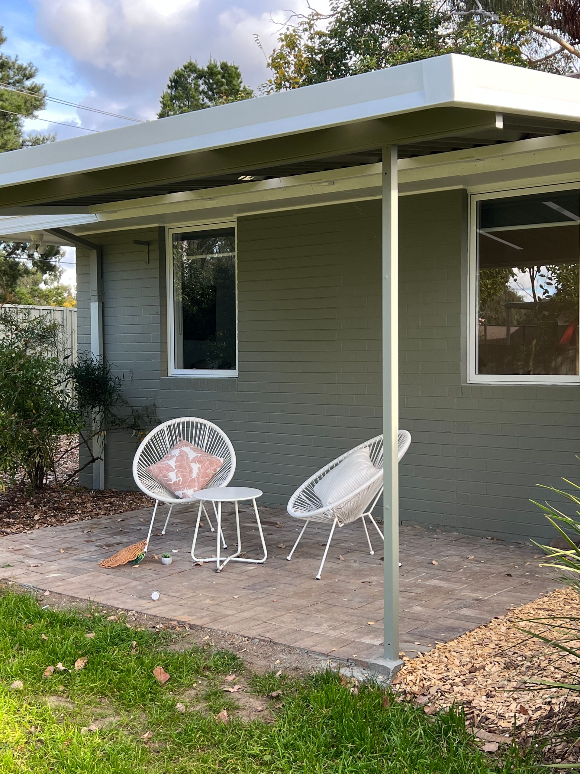 Two white wicker chairs on a concrete patio under a covered area, green building exterior.