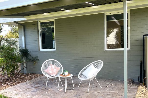 Two white chairs and a small table on a patio next to a painted green brick wall and windows.