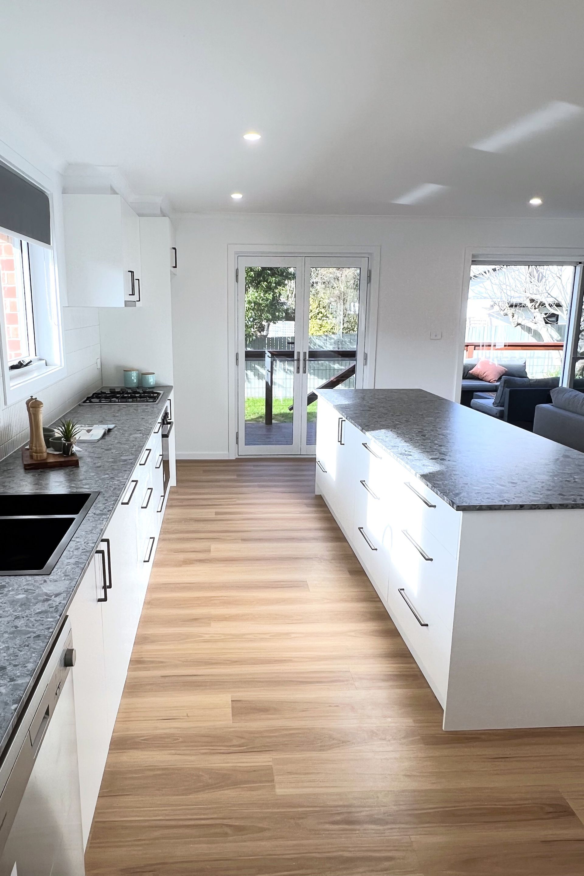 Modern kitchen with white cabinets, wood floors, and a granite countertop island. Sliding glass doors lead outside.