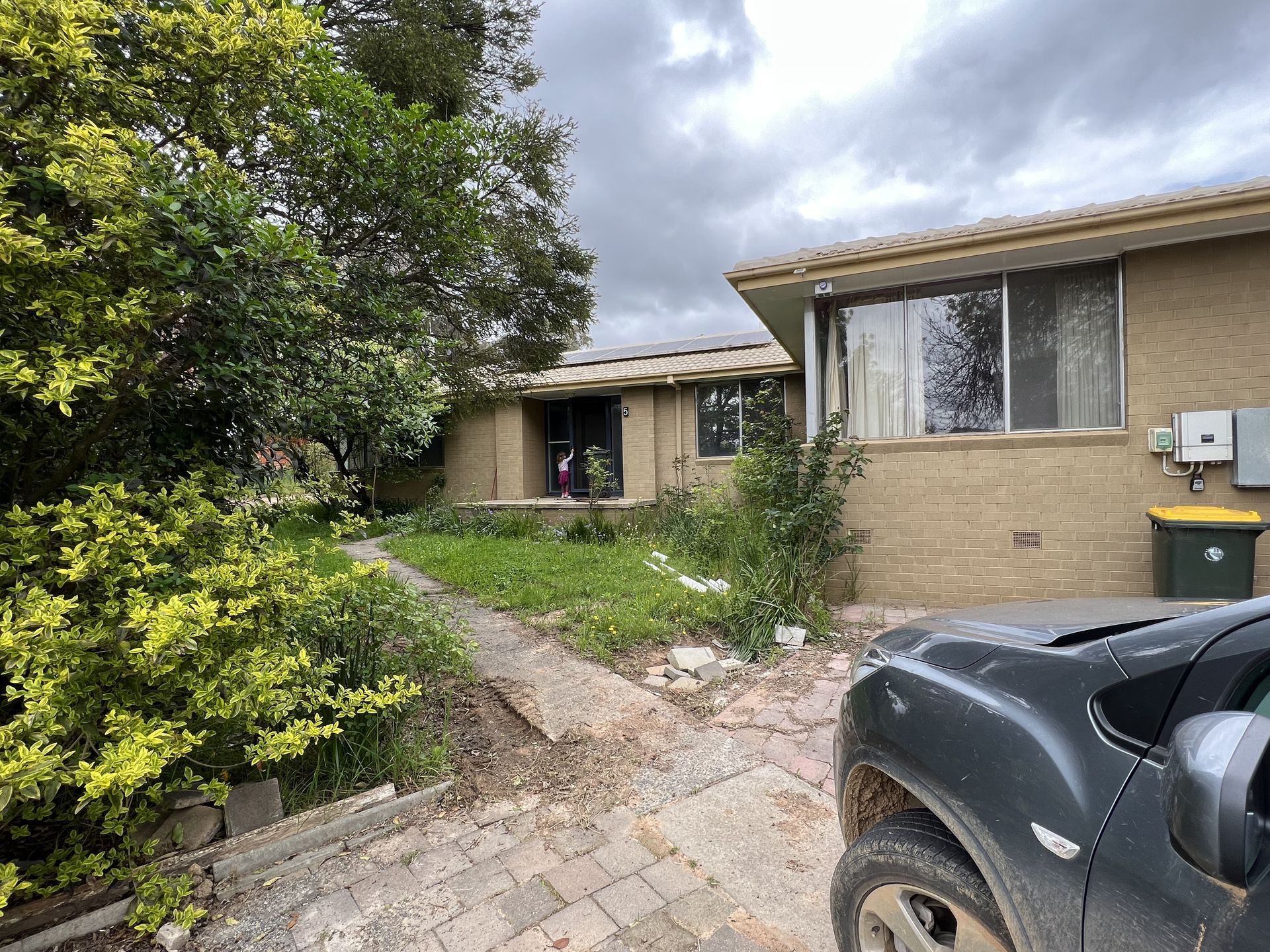 Exterior view of a brick house with a front yard overgrown with weeds. A parked vehicle is in the foreground.