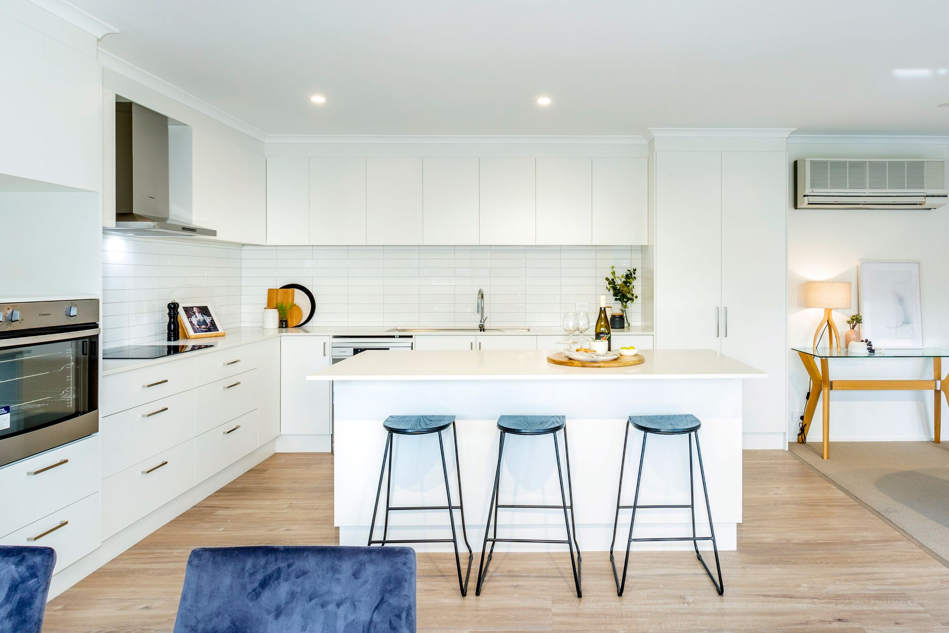 Modern white kitchen with island, stools, and built-in appliances; light wood flooring.