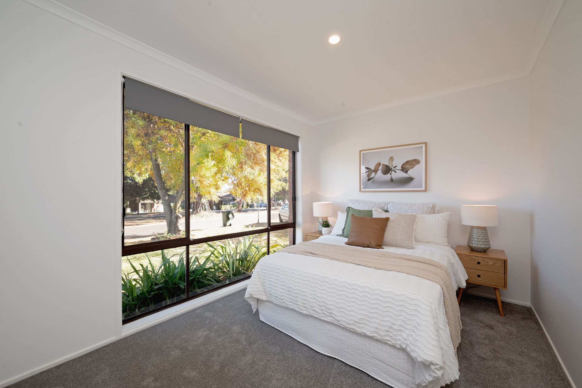 Bedroom with a large window overlooking a park; white walls, gray carpet, bed with neutral bedding, nightstand.