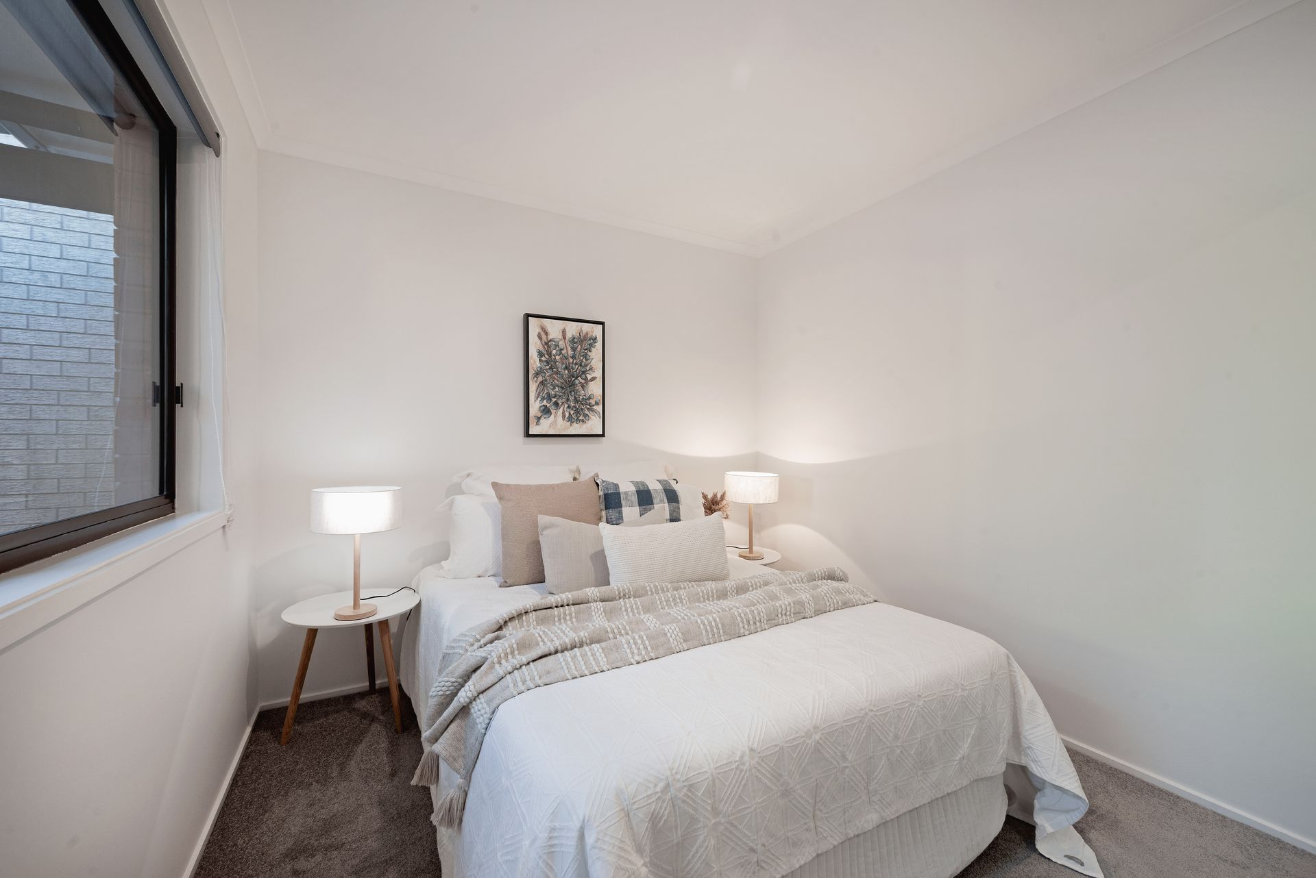 Small, white bedroom with double bed, side table, lamps, and artwork; window on left.