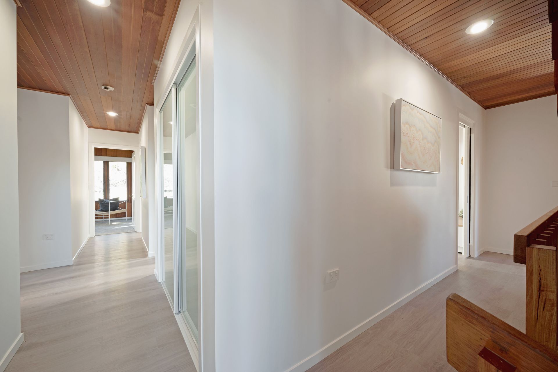 Hallway with wooden ceiling and light-colored floors. White walls with sliding glass door and artwork.