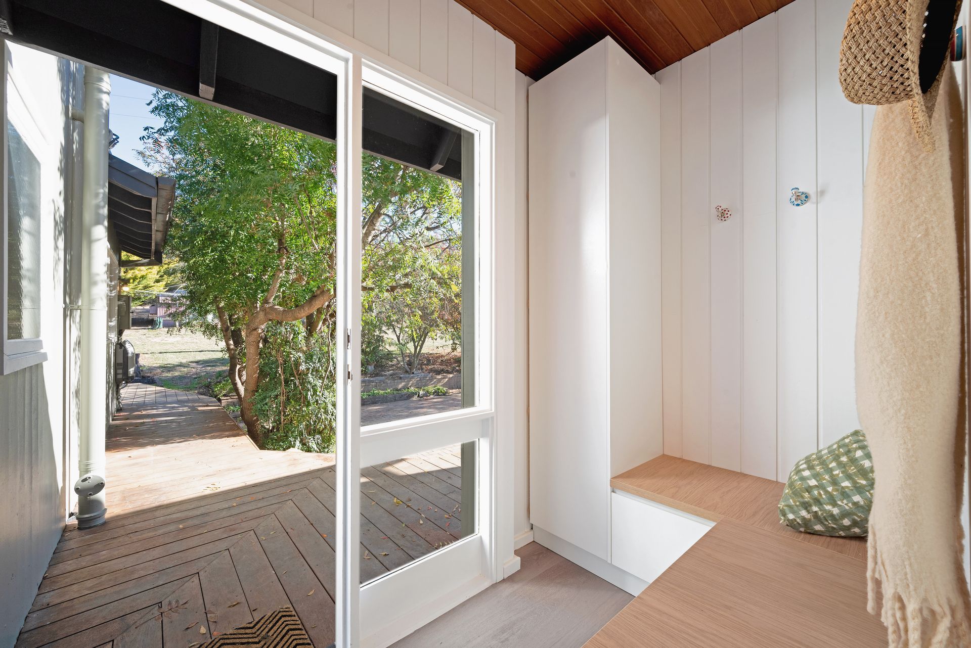 Bright entryway with a glass door overlooking a backyard. White walls and wooden floor. A coat and a green pillow hang.