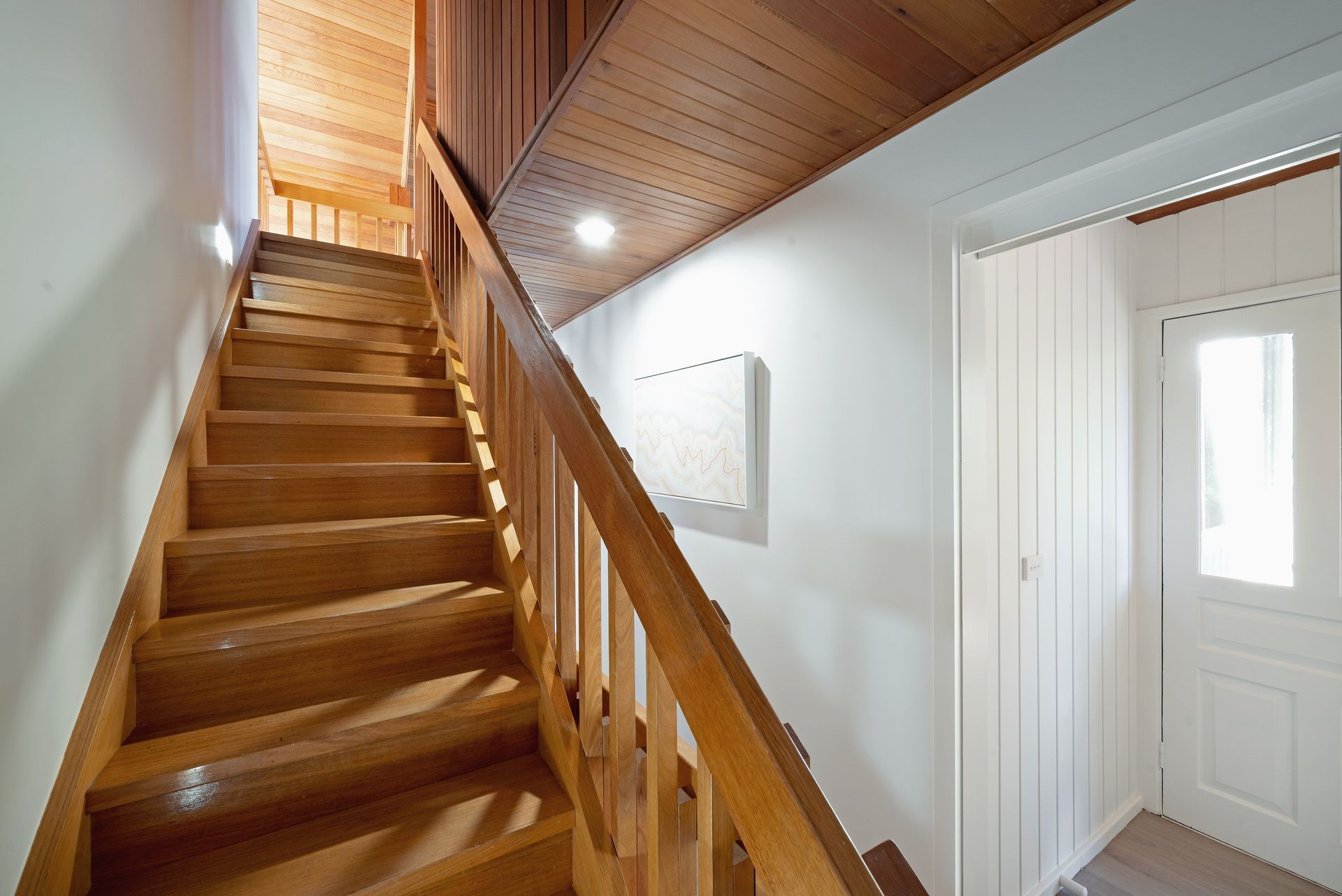 Wooden staircase with wooden banister, leading upwards, lit by recessed lighting. White door on the right.