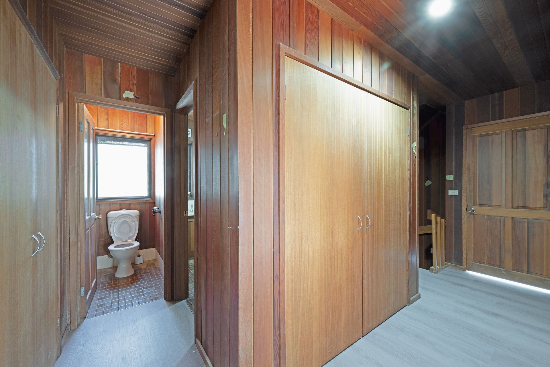 Wooden-paneled hallway with closed closet doors, leading to a bathroom with a toilet and window.