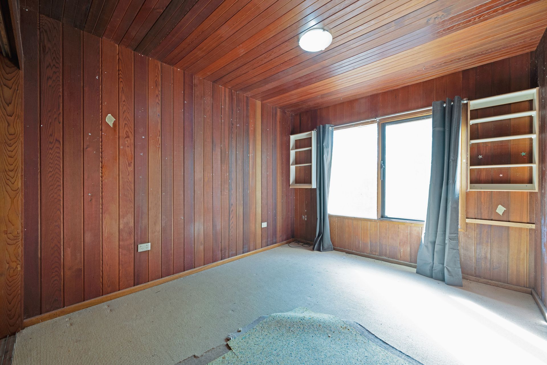 Empty room with wood-paneled walls and ceiling, a window with curtains, built-in shelves, and carpet.
