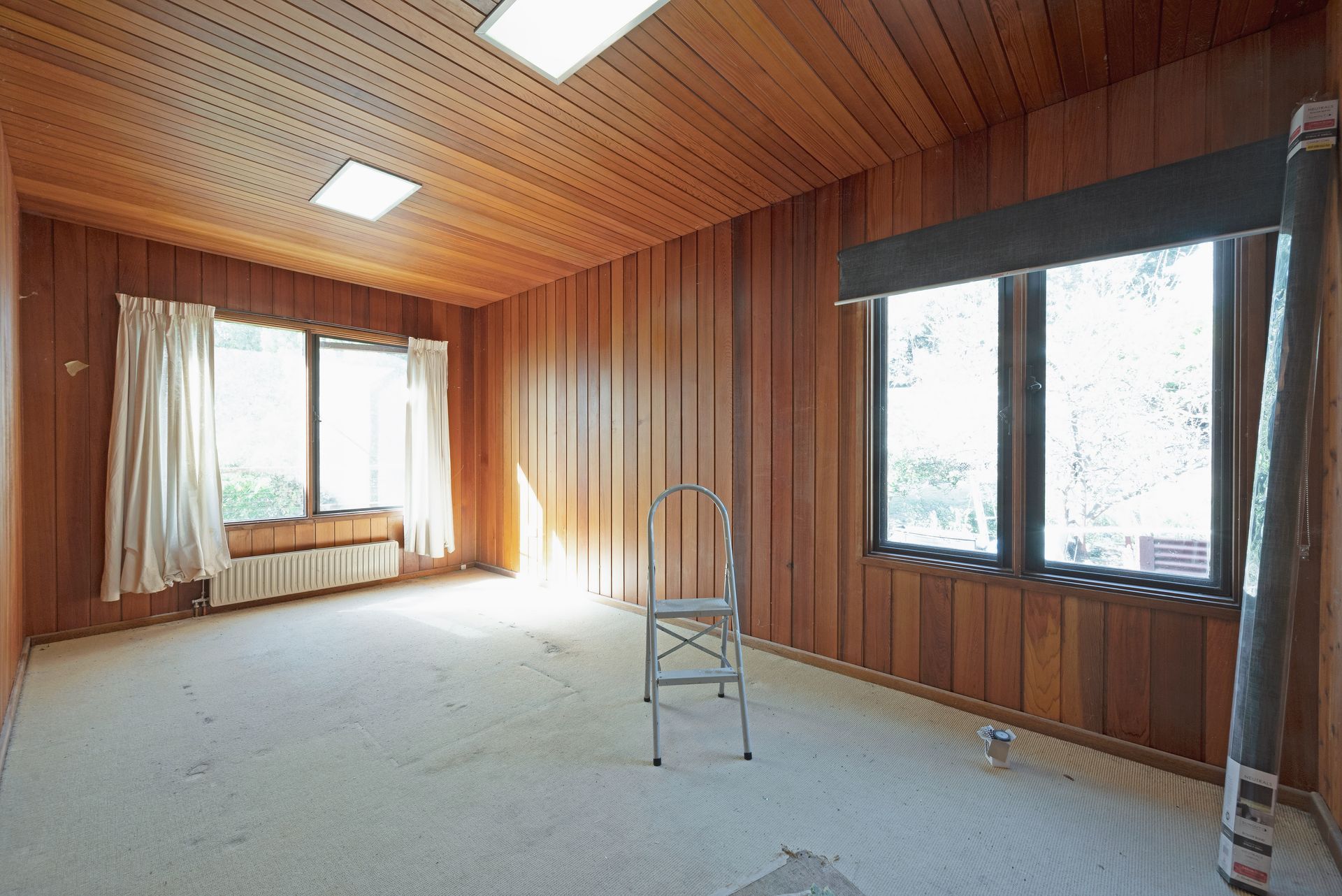 Empty room with wood paneling on walls and ceiling, two windows, and a metal step stool.