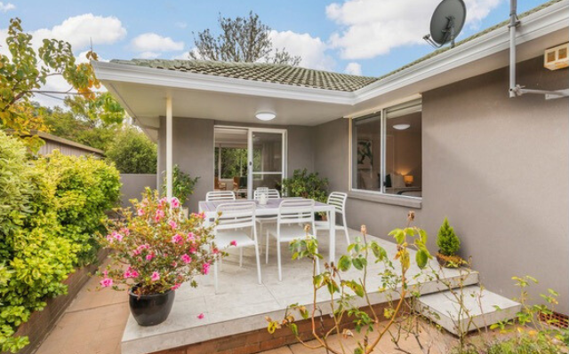 Patio with white table and chairs, potted flowers, and a gray house under a blue sky.