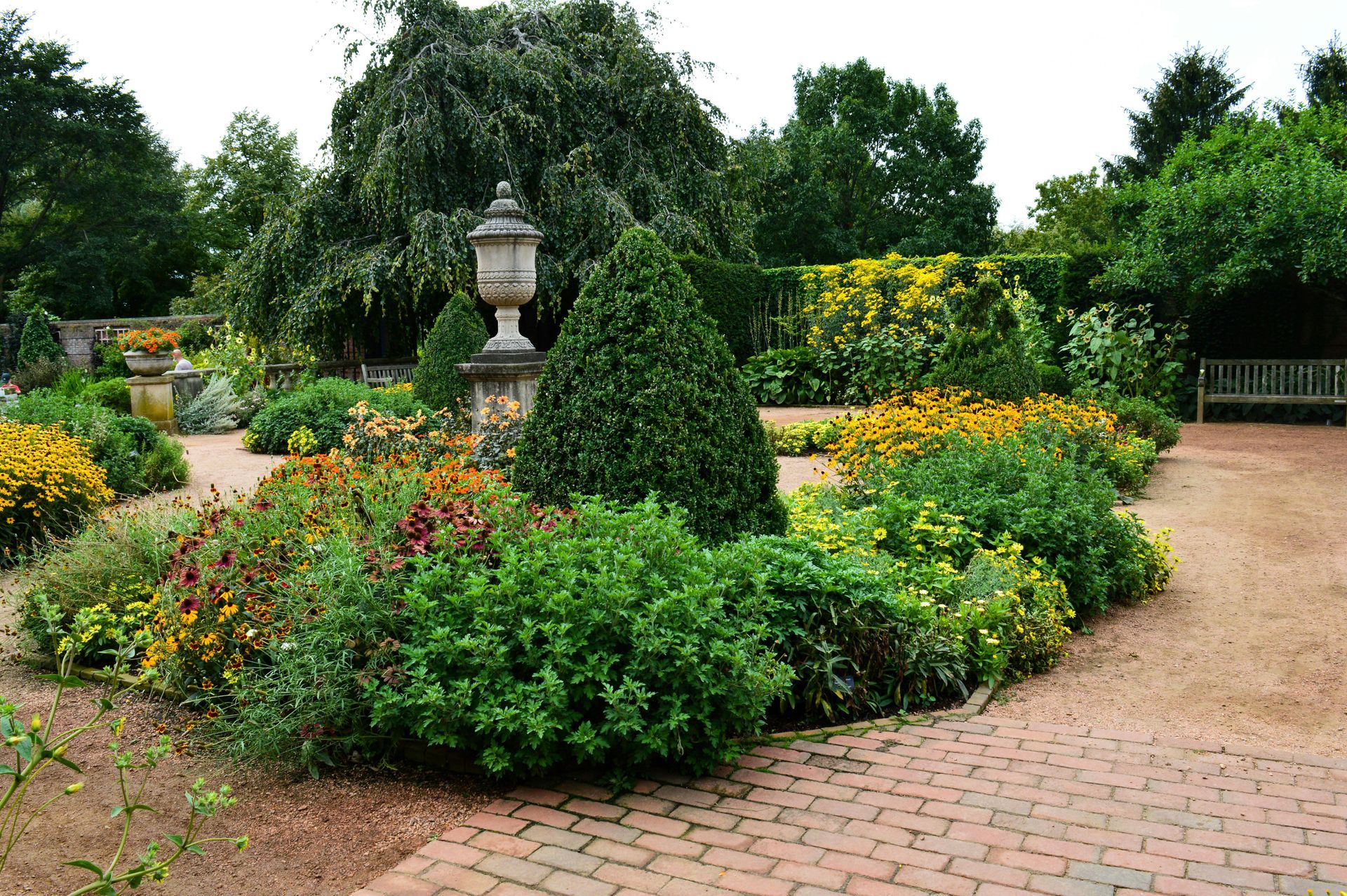 Garden with brick pathways, flower beds, and an urn statue. Lush greenery and colorful blooms.