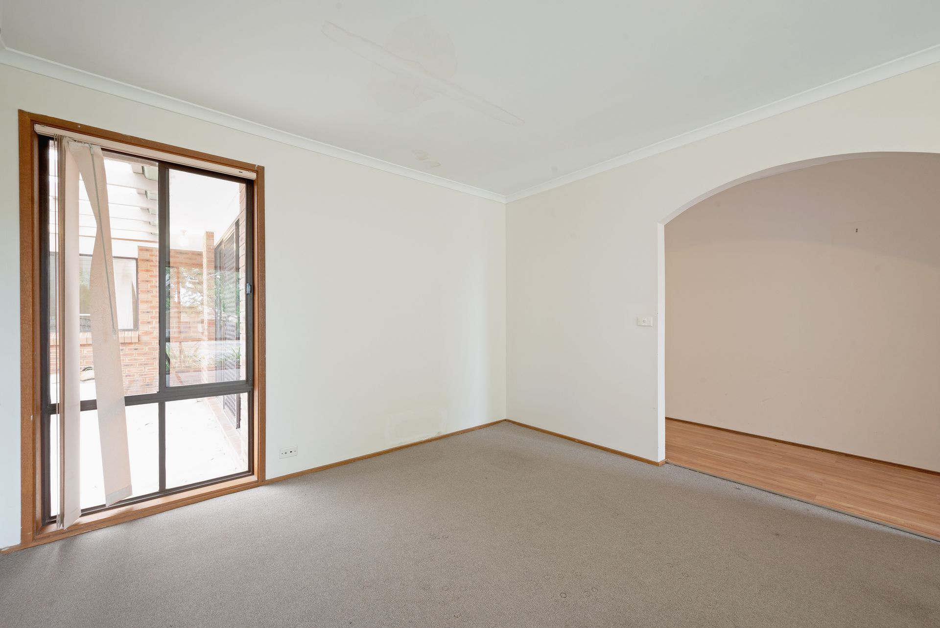 Empty room with brown carpet, window with curtains, and arched doorway to another room.