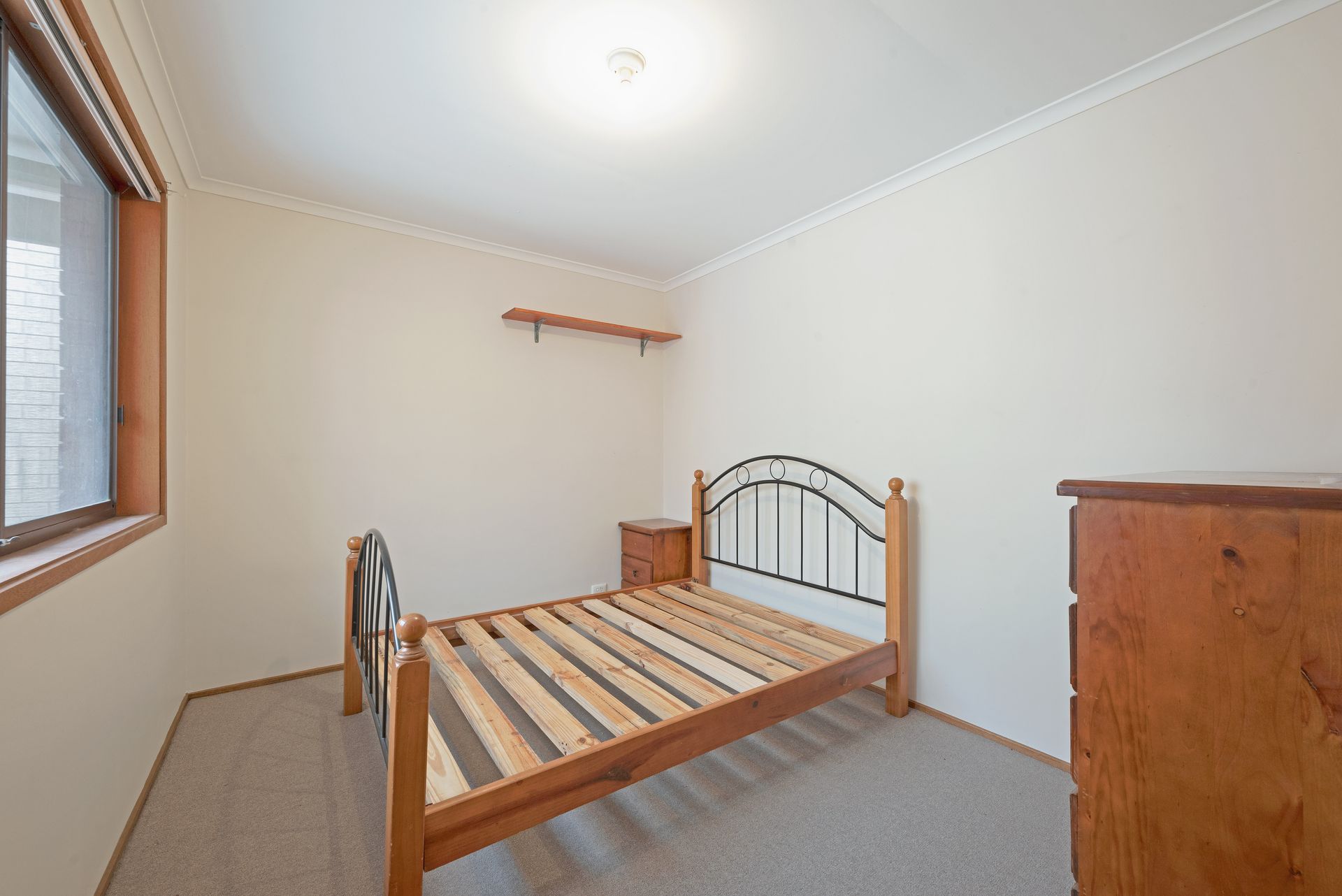Bedroom with wooden bed frame, nightstand, dresser, and window. Walls are white, carpet is gray.
