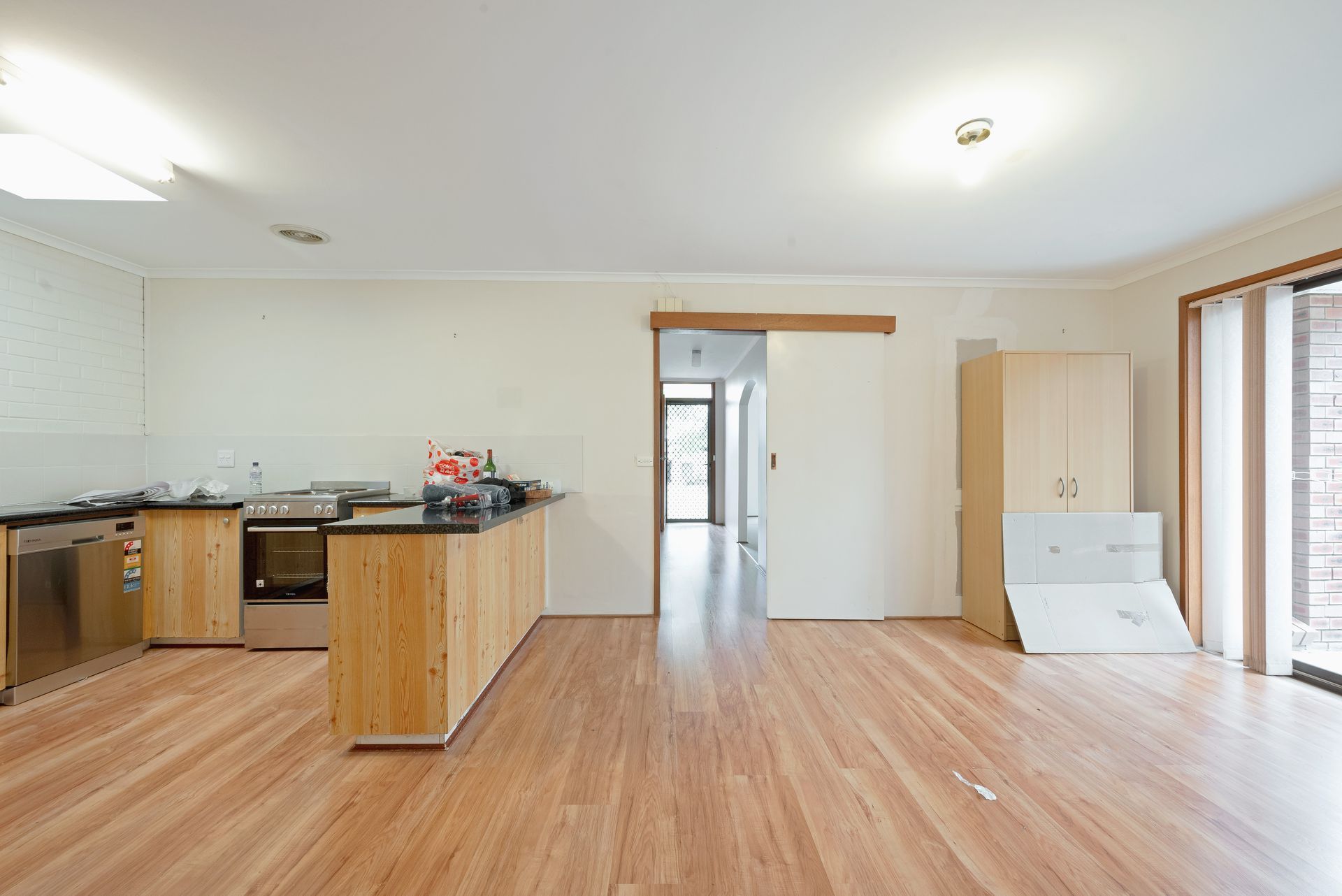Interior shot of a kitchen and living space with wooden floors, light walls, and a sliding door leading to a hallway.