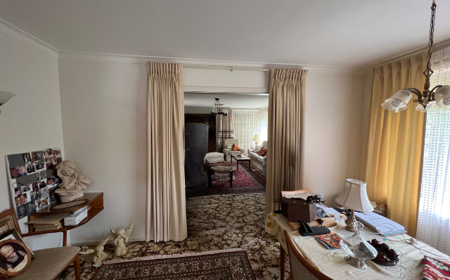 Interior view of a room with beige curtains framing a doorway. A cluttered table and a rug are in the foreground.