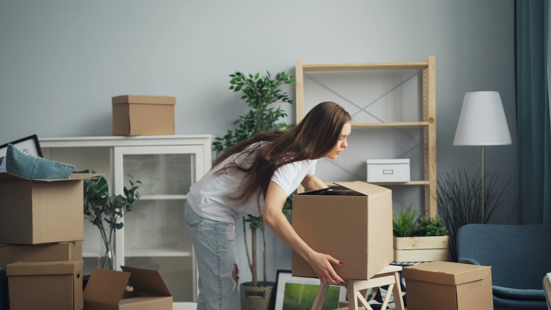 Woman lifting a cardboard box in a room with other boxes, furniture, and plants.