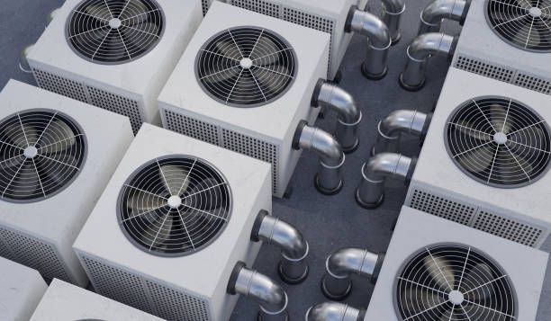 Rows of white air conditioning units with fans and metal pipes on a rooftop.