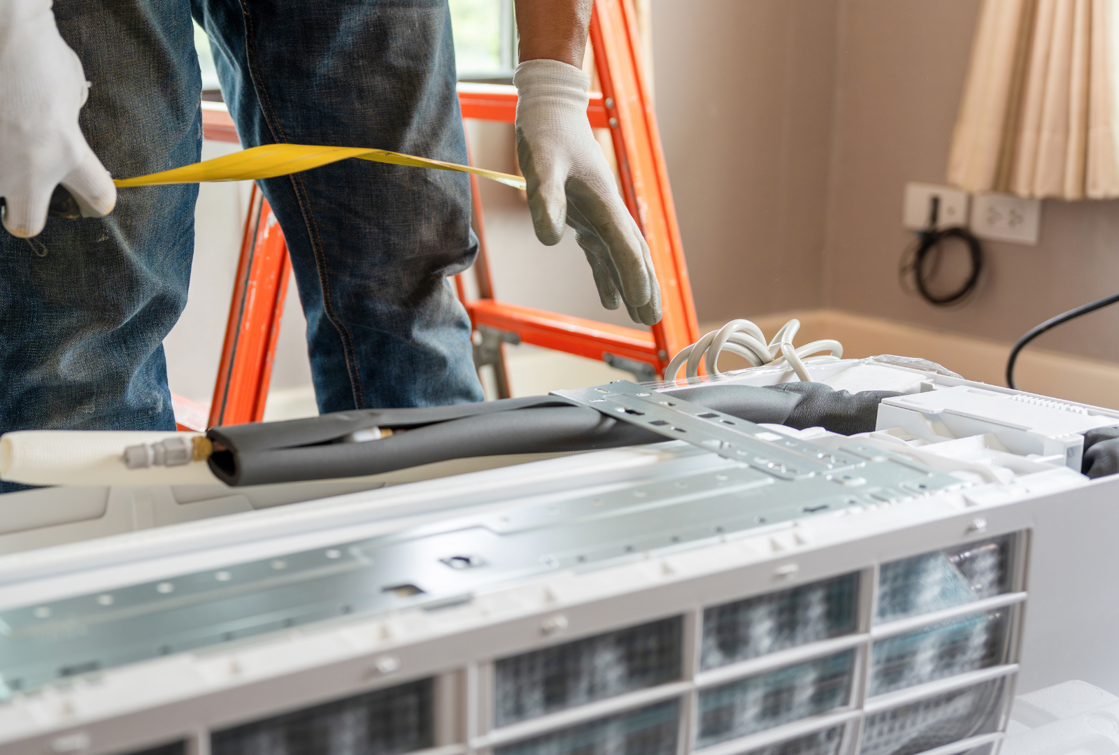 Person in gloves installing AC unit indoors near a ladder, holding a yellow measuring tape.