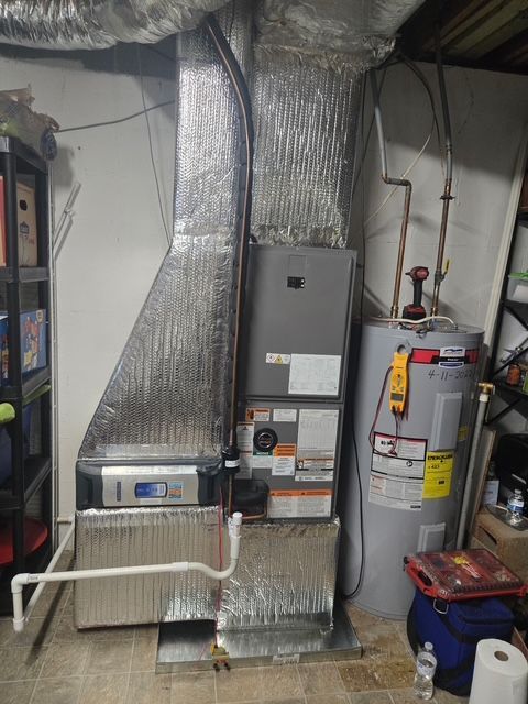 Furnace and water heater in a basement. Metal ducts and insulation visible. Tools are nearby.