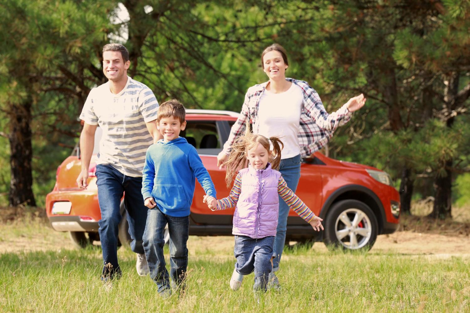 Family of four running toward camera outdoors near a red car, smiling.