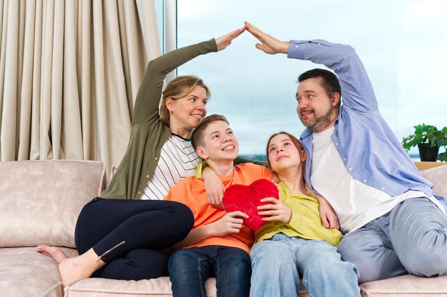 Family of four on a couch, holding a heart; parents make a roof with arms, looking up.
