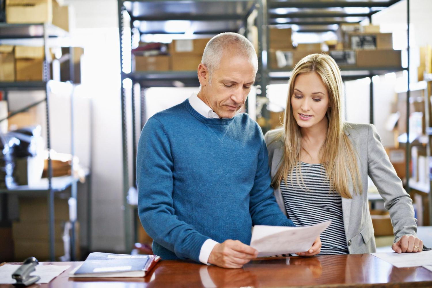 Man and woman looking at paperwork in a warehouse setting, surrounded by boxes on shelves.