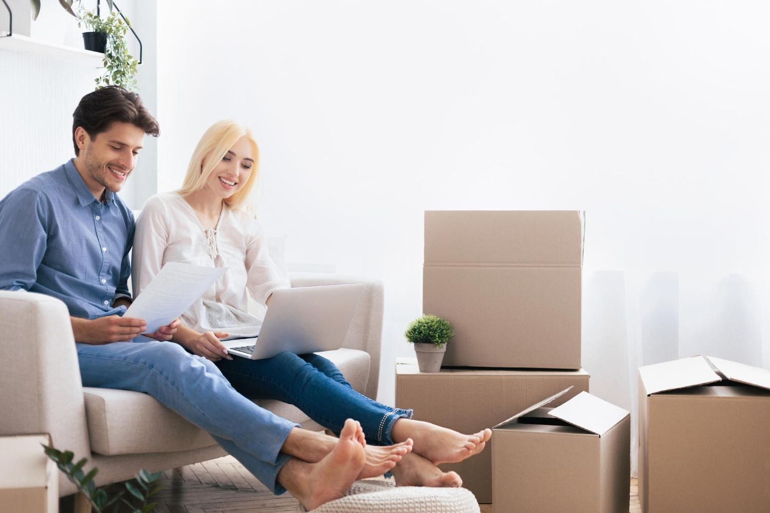 Couple sitting in new home with boxes, using a laptop and paperwork, smiling.