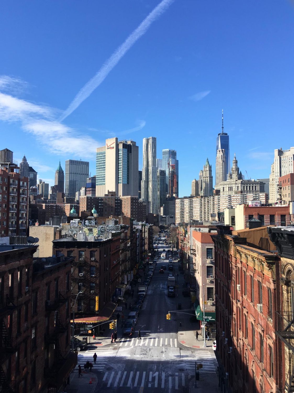 City street view with skyscrapers in the distance, clear blue sky, and a contrail.