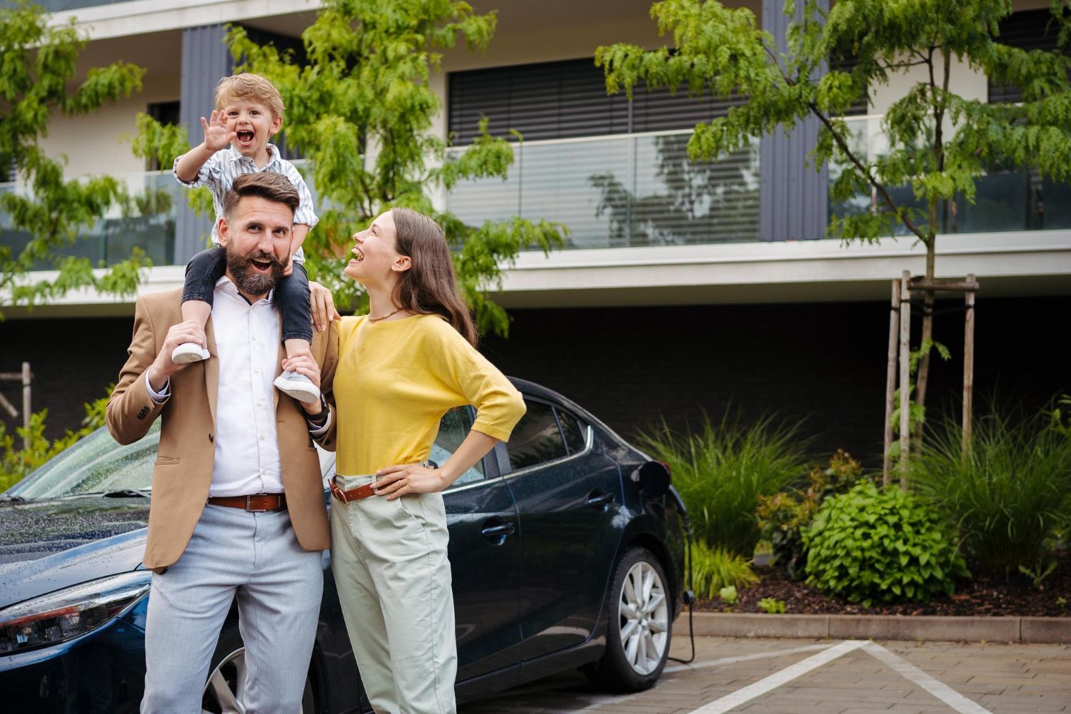 Family near a car; father with child on shoulders, mother smiles. Modern building in background.