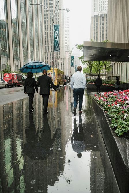 People walking on a wet city street, one with a blue umbrella, reflecting in puddles.