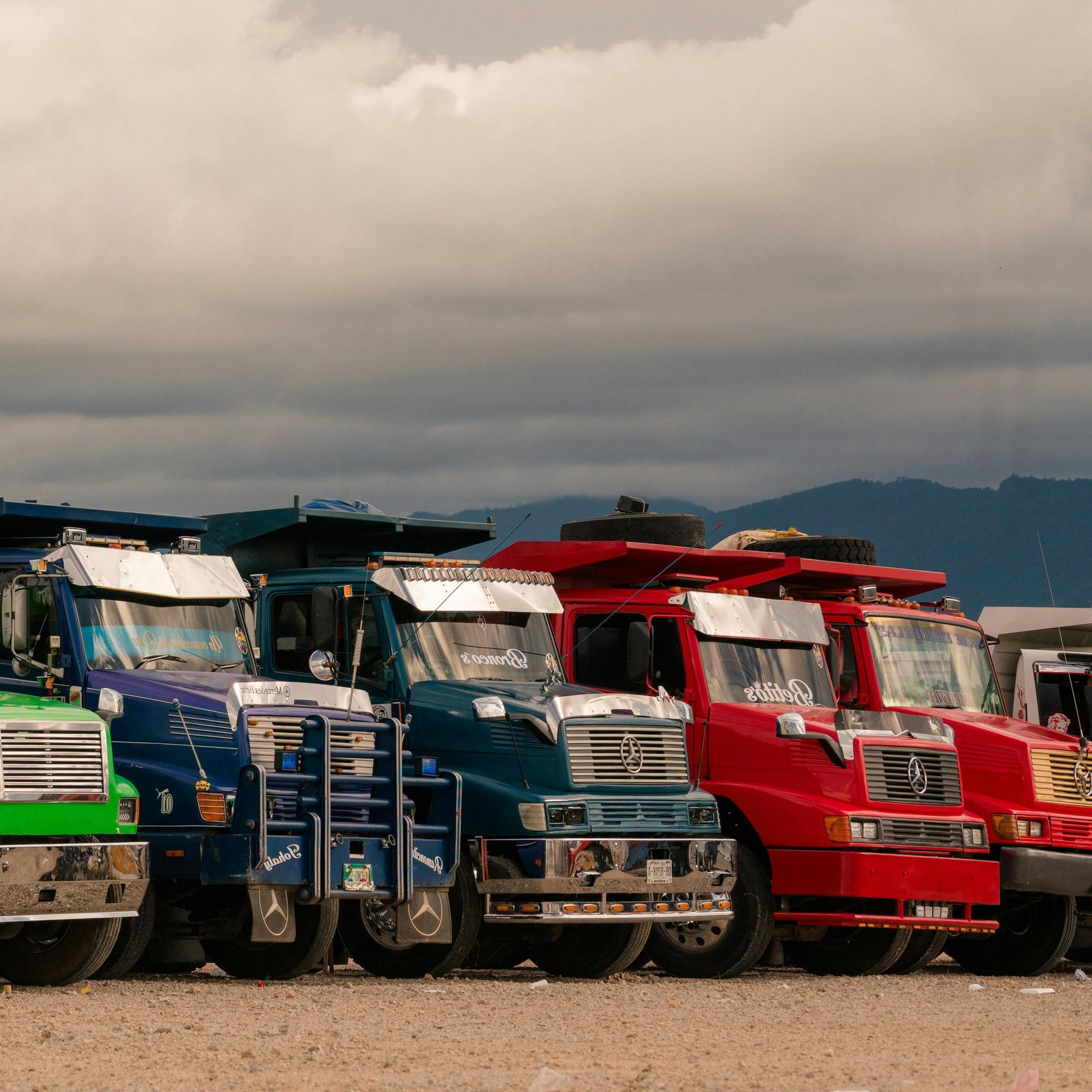 Colorful dump trucks parked in a row under a cloudy sky, mountains in the background.
