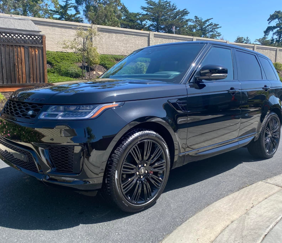 Black SUV parked on a sunny street beside a brick wall and wooden fence