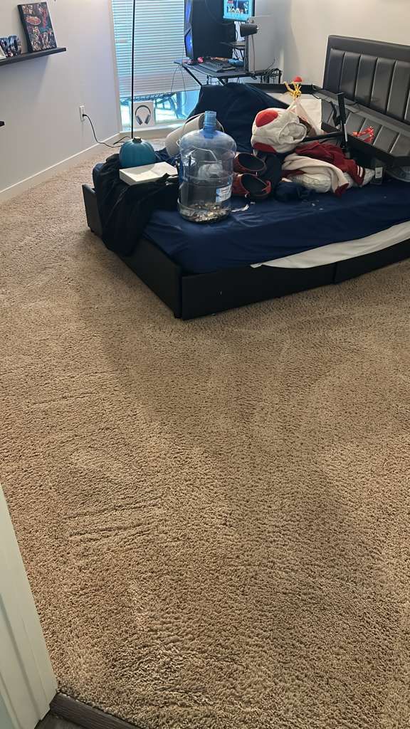 A messy bed with a large water jug sits in a room with tan carpet and a small wall shelf.