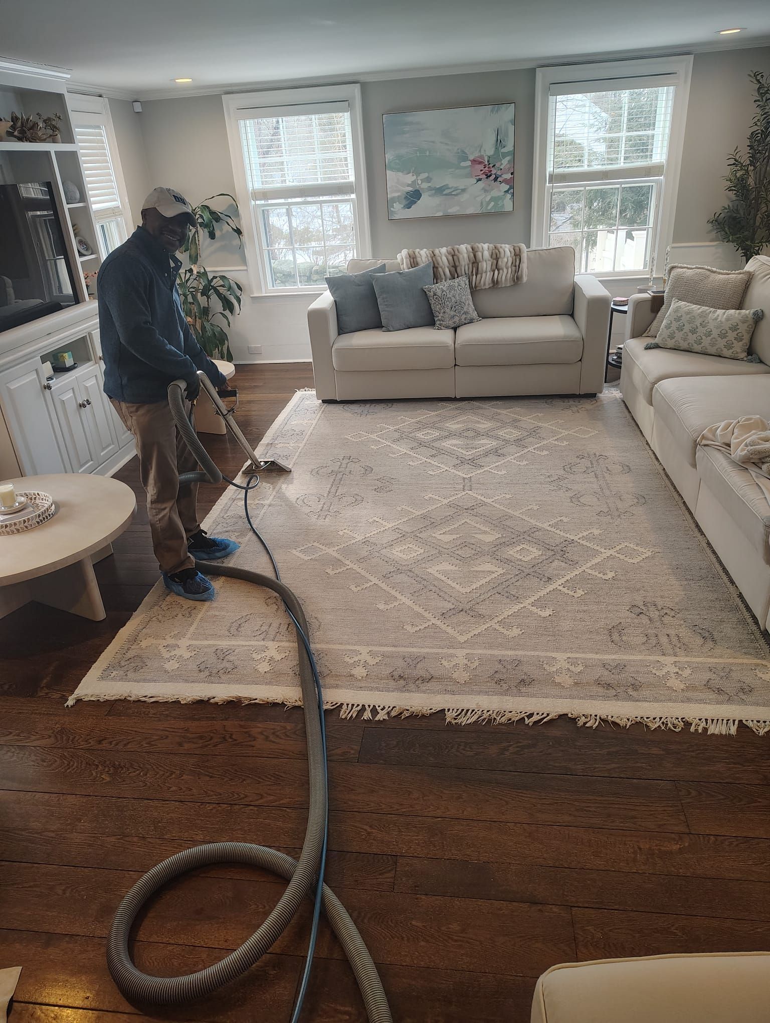 A person in a baseball cap uses a professional vacuum wand to clean a large, patterned rug in a bright, furnished living room.