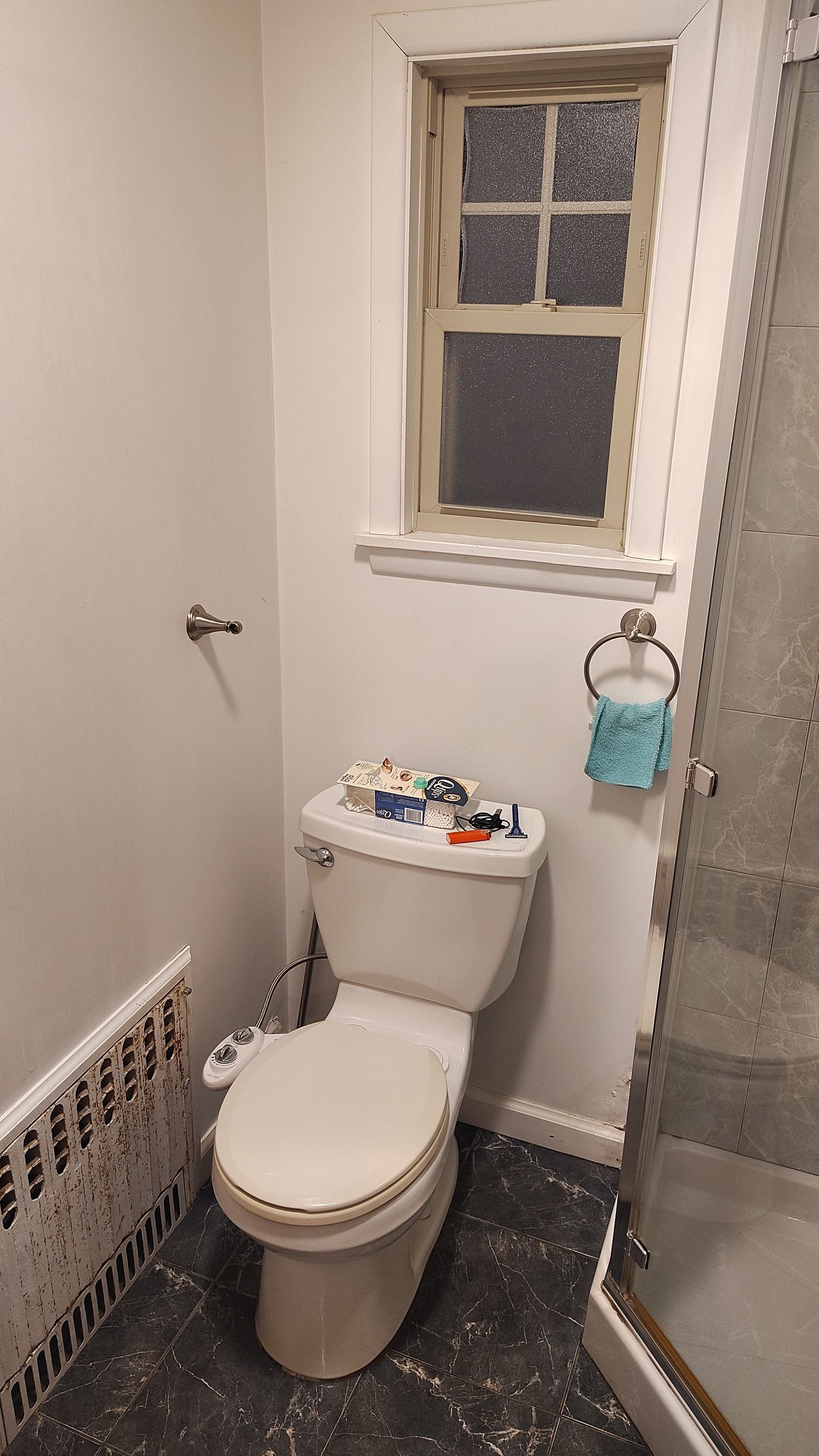 A white toilet with a bidet attachment in a bathroom with dark flooring, a window, and a shower enclosure.