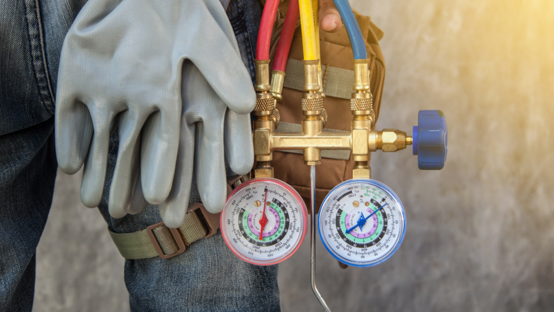 Gloved hand beside a brass manifold with colored hoses and two pressure gauges against a tan backdrop