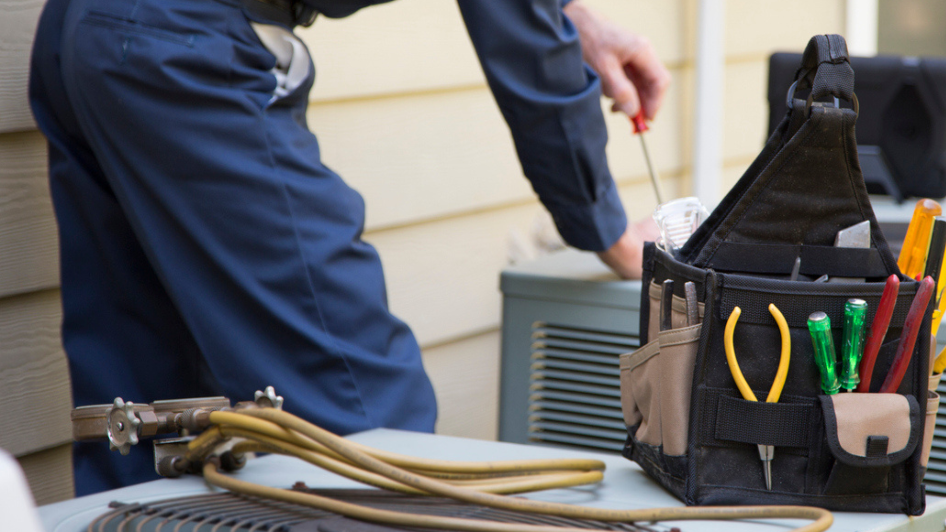 Technician kneeling by outdoor HVAC unit with tool bag and hoses beside a house wall