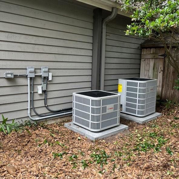 Two air conditioning units on concrete pads next to a house with gray siding.