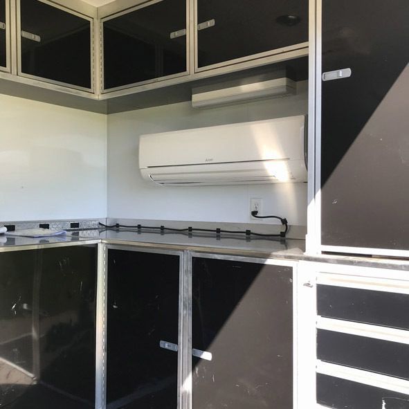Interior of a trailer with an air conditioning unit mounted on a white wall. Black cabinets below and above.