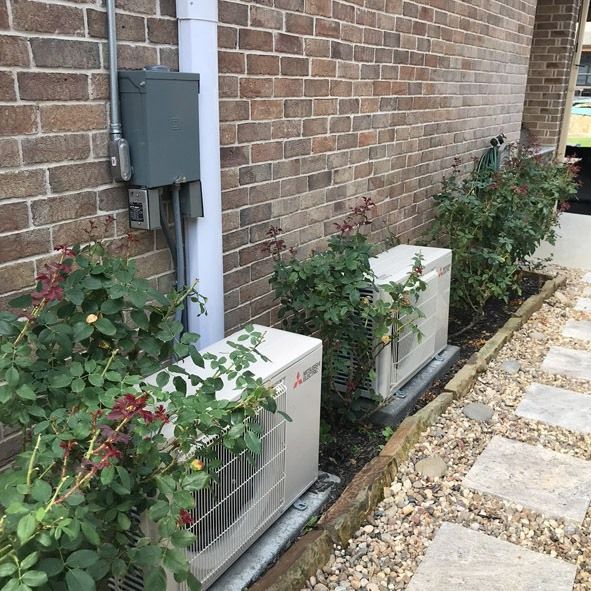 Two white HVAC units sit next to rose bushes and a brick wall. A pathway runs alongside.