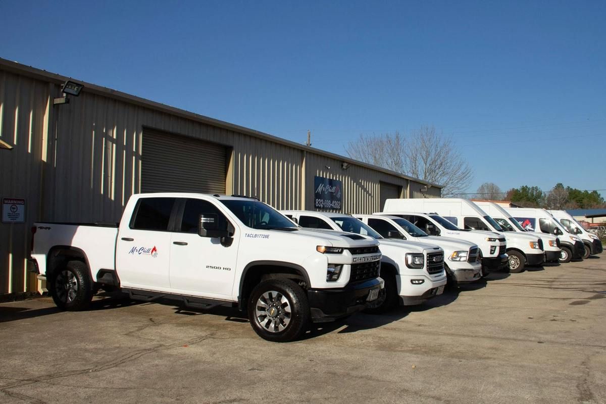 White trucks and vans parked in a row in front of a tan industrial building on a sunny day.
