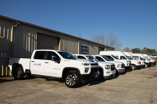 White trucks and vans parked in a row in front of a tan industrial building on a sunny day.