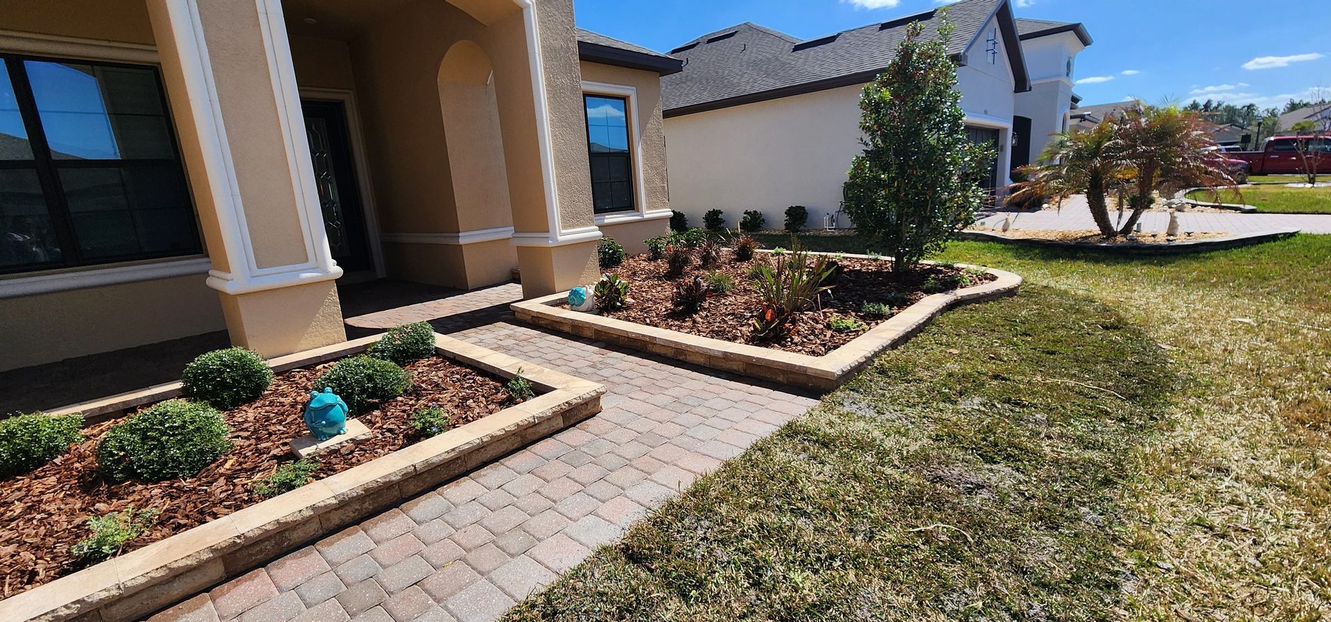A brick walkway leads to a house with a beige facade and lush landscaping. Blue sky overhead.