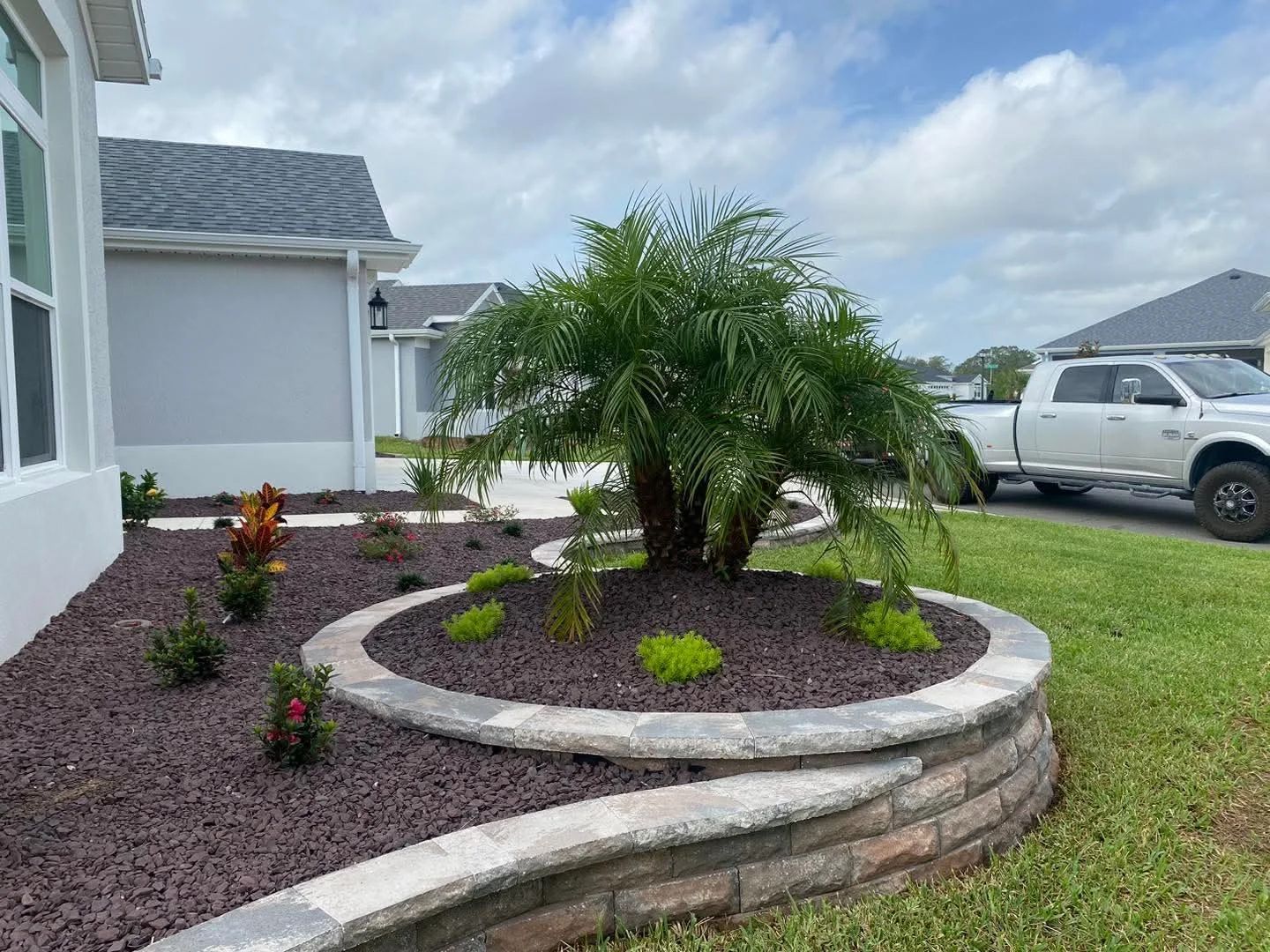 Landscaped yard with palm tree in raised bed, bordered by stone wall, next to a house and truck.